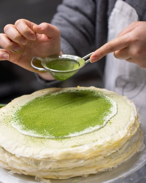 Angeschnittener Mille Crêpe Cake, über den Matcha gesiebt wird — die einzelnen Crêpe-Lagen sind sichtbar